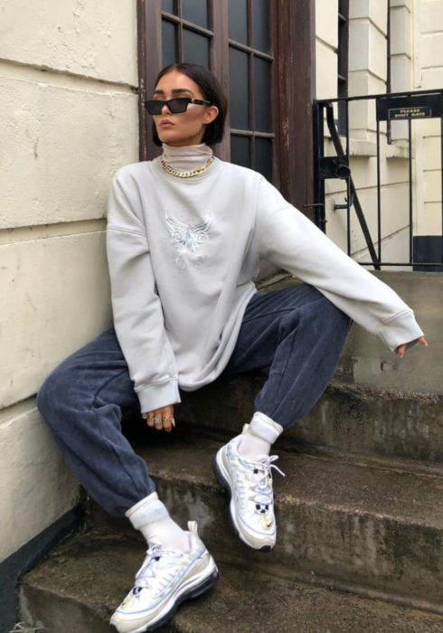 Looks con sudadera; mujer con lentes de sol sentada en escaleras, con hoodie oversized gris blanco, mom jeans y tenis deportivos