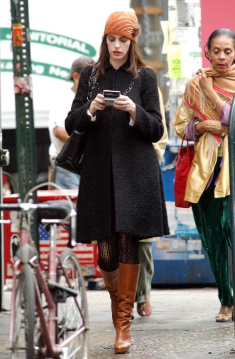 Escena de la película El Diablo Viste a la moda. Andrea caminando por la calle mientras usa un abrigo negro, botas y un sombrero de color naranja