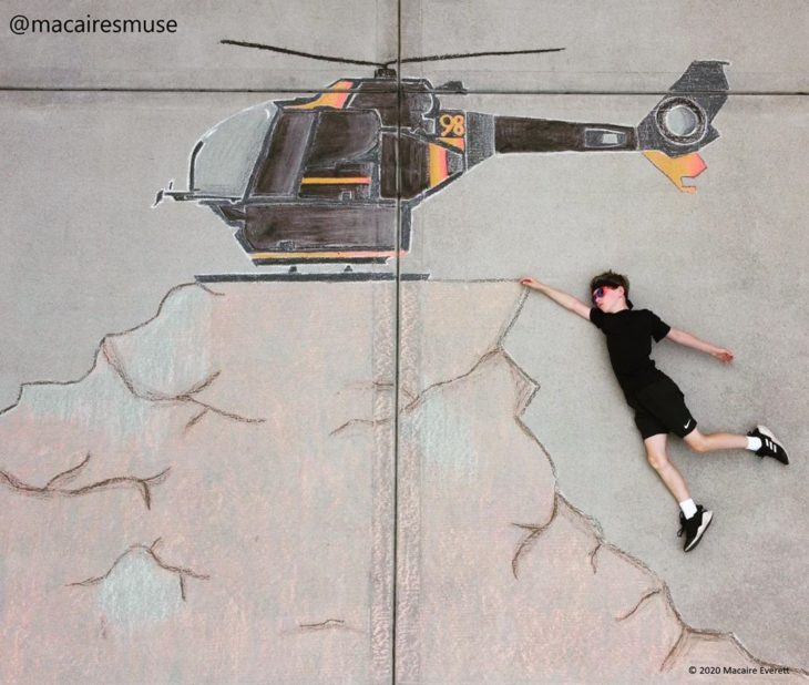 Dibujo de hecho con tiza de un helicoptero negro sobrevolando una montaña, niño recostado como si se sostuviera de la montaña