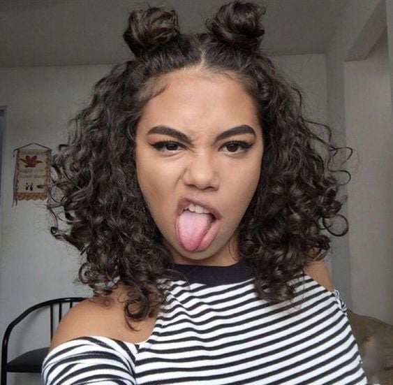 Chica morena de cabello rizado con blusa de rayas sacando la lengua con dos chonguitos en la cabeza
