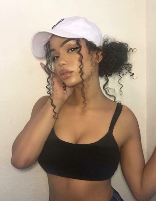 Chica morena de cabello rizado con una gorra blanca en la cabeza y un chongo