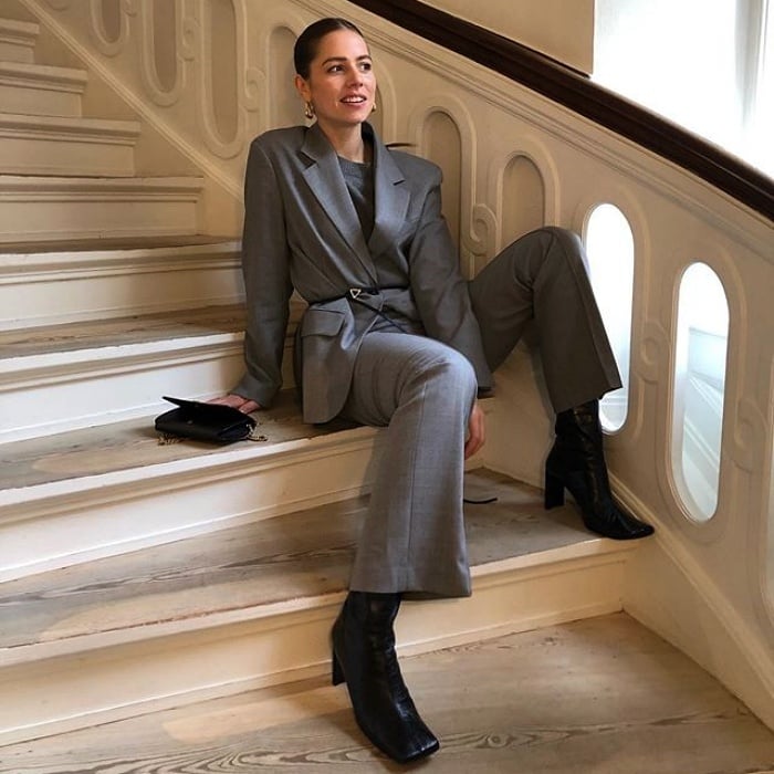 chica de cabello castaño con un saco gris, top gris, pantalones de vestir gris y botas negras de cuero