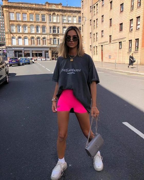 Chica rubia caminando por la calle con blusa gris larga y pantaloncillos de ciclista rosa neón