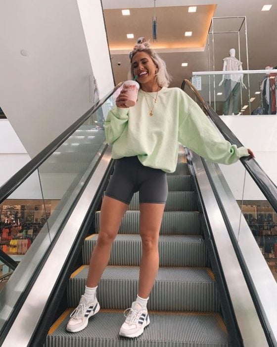 Chica rubia en escaleras eléctricas sosteniendo un café mientras usa sudadera verde pistache y shorts de ciclista grises con tenia blancos