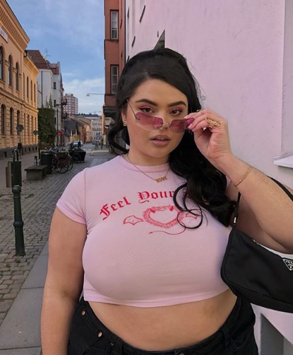 chica curvy de cabello castaño largo usando lentes de sol rosa, camisa crop top rosa con letras al frente, pantalón negro y bolso de mano de piel