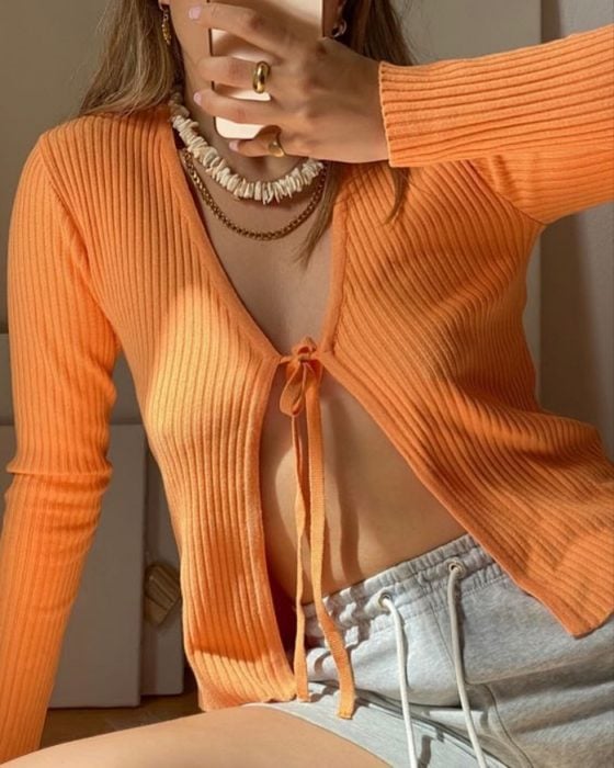 chica tomandose un selfie usando un cardigan tejido color naranja claro o suave usando collares de perlas y de oro