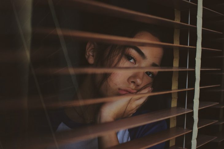 Chica mirando por la ventana, detrás de una persiana