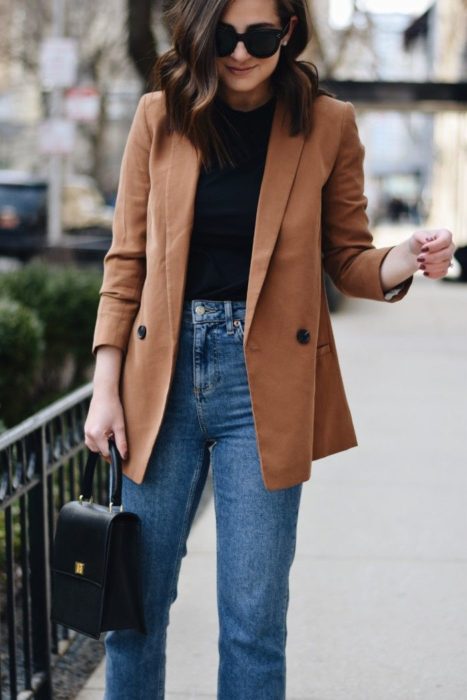 Chica usando blazer color camel, con suéter negro y jeans denim, además de bolsa de mano negra y lentes oscuros