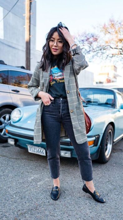 Chica usando blazer de color gris con estampado a cuadros, con playera negra de estampado, y zapatos y pantalón negros