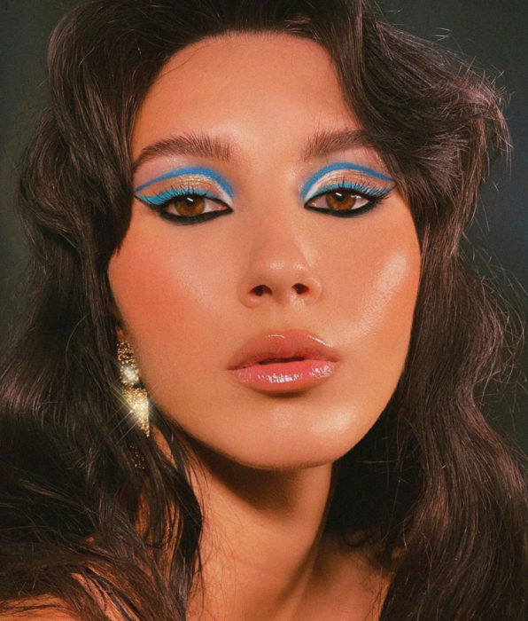 Mujer morena de cabello castaño oscuro ondulado, ojos café, delineado gráfico color negro y azul cielo, labios nude