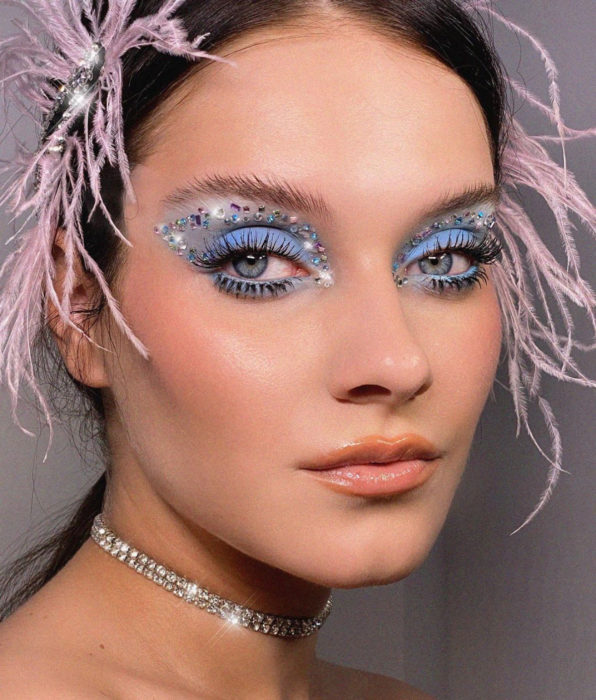 Mujer con maquillaje para fiesta, ojos azules, delineado gráfico con brillos glitter, sombra azul cielo, labios nude, cabello castaño oscuro, peinado con tocado de plumas rosas