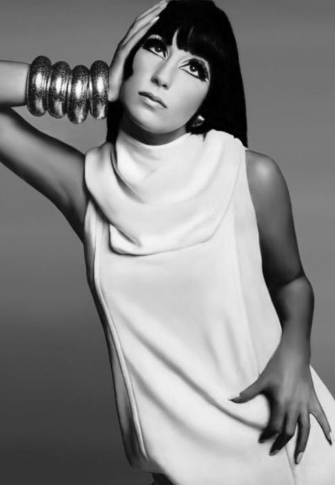 Looks icónicos de Cher; cabello negro, lacio, largo con fleco, pestañas postizas, vestido blanco, pulseras grandes