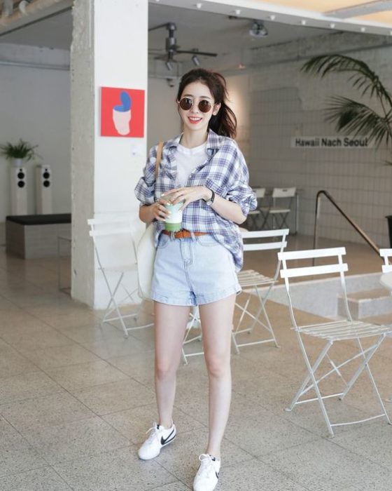 Chica asiática de cabello largo en una coleta usa lentes de sol, blusa blanca, camisa de cuadros, shorts de mezclilla y tenis blancos
