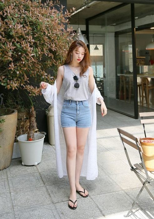 Chica asiática con cabello suelto claro con blusa gris de manga corta, fajada en shorts de mezclilla y kimono blanco