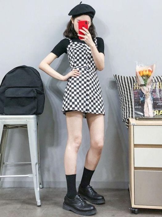 Chica asiática se toma selfie frente al espejo con vestido corto blanco con negro y blusa negra por debajo con botas negras y gorro