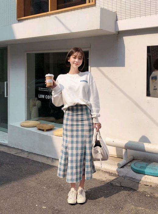Chica asiática de cabello corto con sudadera blanca y falsa larga de cuadros y sostiene un café