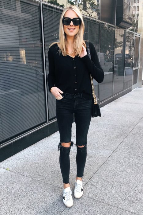 Chica usando jaens rasgados, blusa de manga larga en color negro y tenis blancos, además de lentes oscuros y bolso negro