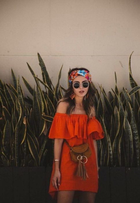 Chica usando outfit veraniego de vestido naranja off the shoulder, arriba de la rodilla, con lentes oscuros banda en el cabello y bolso cruzado