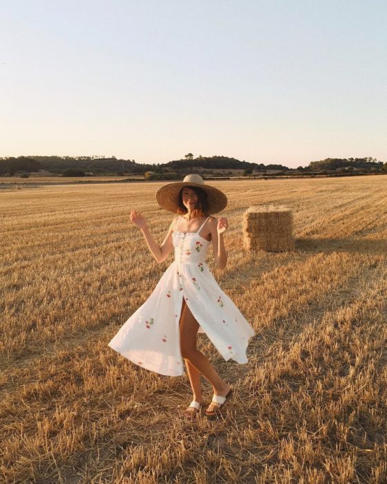 Chica usando outfit veraniego de vestido con vuelo, color blanco y estampado, y abertura de un lado y sombrero de paja