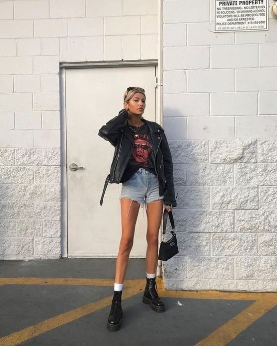 Chica rubia con cabello recogido posa en la calle con chaqueta negra, short de mezclilla y botas dr. martens