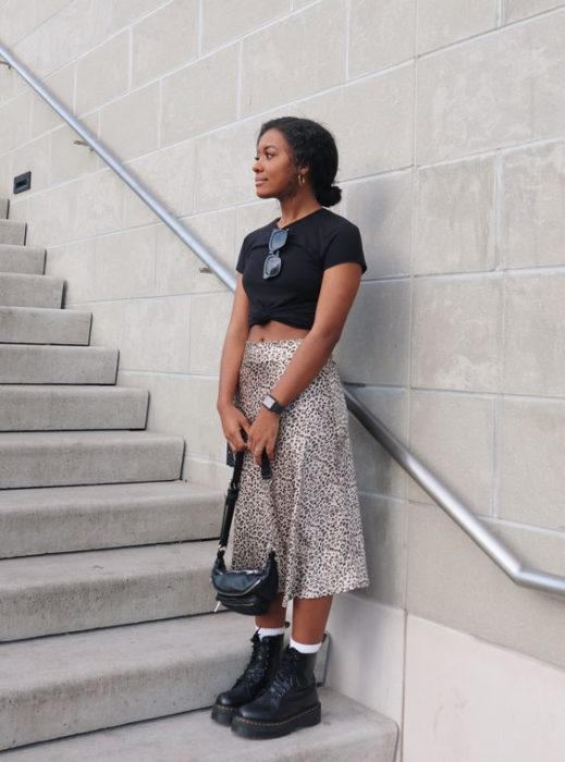 Chica morena con blusa negra y falda de leopard print sosteniendo un bolso negro y usando botas dr. martens