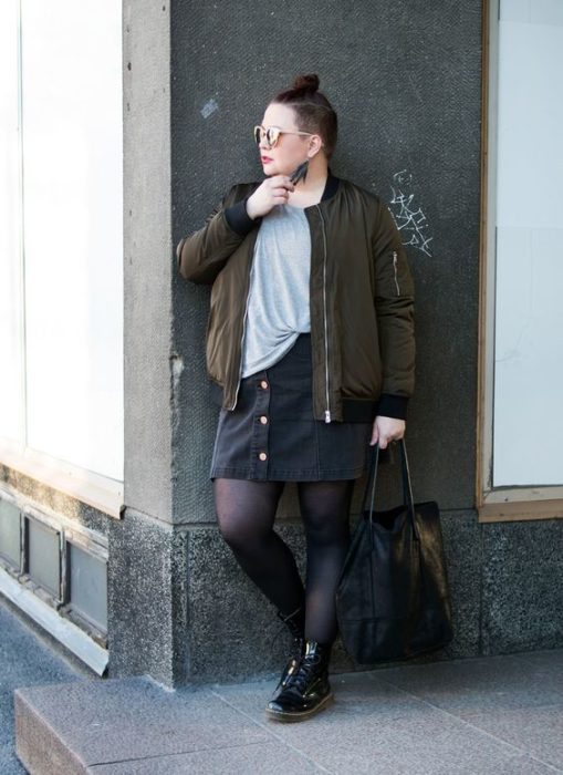 Chica plus size recargada en la pared con chaqueta verde militar, blusa gris, falda negra, medias negras y botas dr. martnes