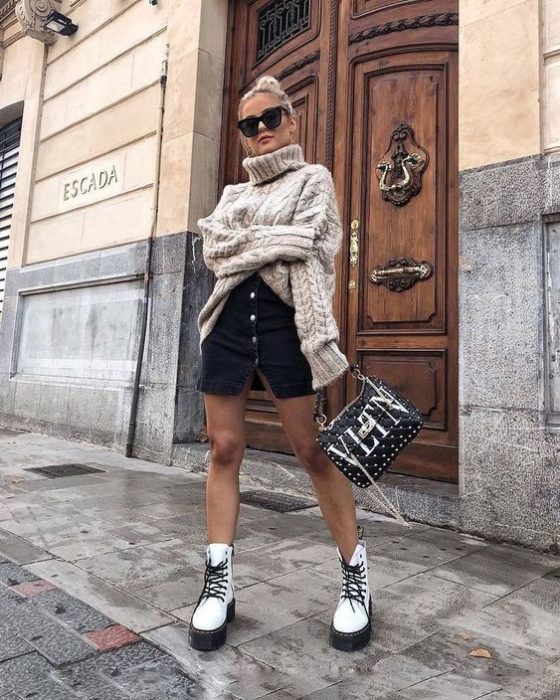 Chica rubia de cabello agarrado en un chongo con lentes de sol negros y suéter oversized beige, falda negra con botones en la parte de adelante y botas dr. martens blancas