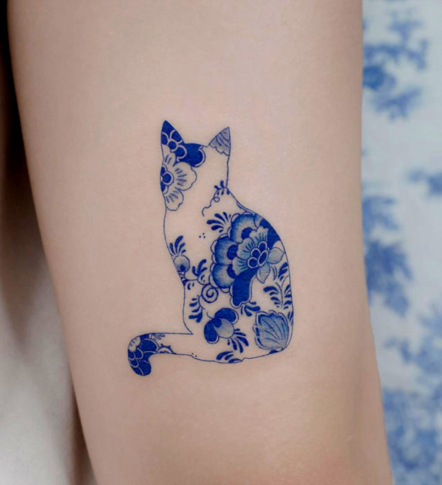 Tatuaje pequeño estilo japonés de tinta azul en el brazo; silueta de gato con flores dentro