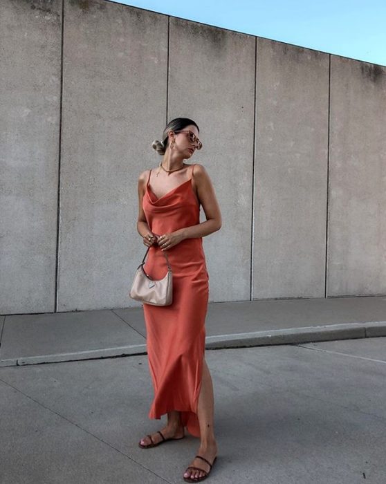 chica rubia con lentes de sol usando un vestido anaranjado de satén slip dress con bolso de mano color beige y sandalias de piso