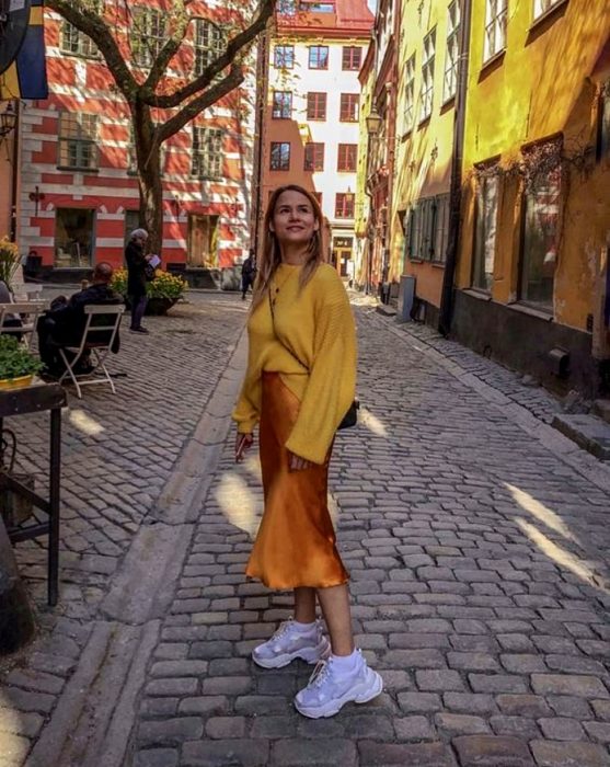 chica rubia usando un suéter amarillo oversized, vestido de satén slip dress naranja con tenis deportivos blancos