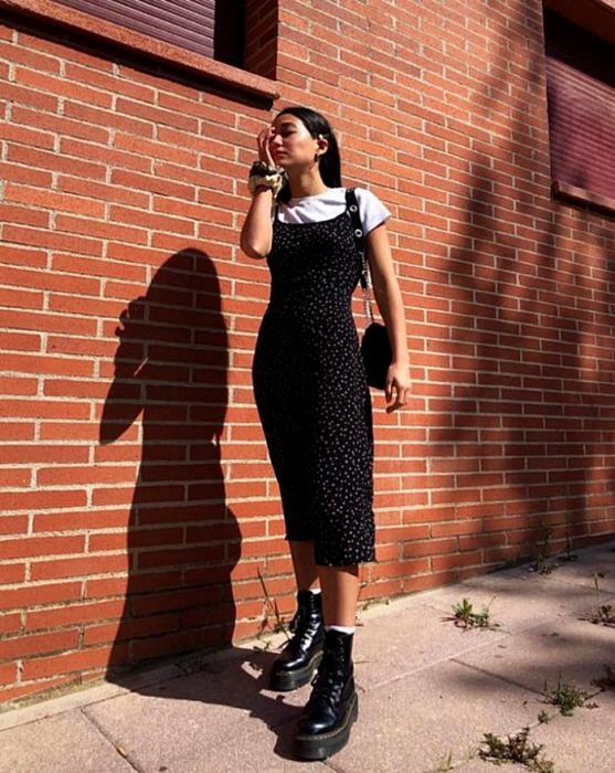 chica de cabello castaño usando un vestido negro de satén slip dress con flores, camiseta blanca de manga corta y botas de cuero negras militares y bolso de mano negro