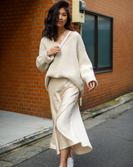 chica de cabello oscuro usando un suéter blanco holgado con vestido de satén color crema y tenis deportivos blancos