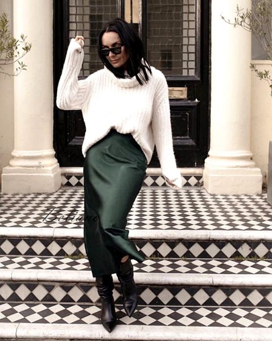 chica de cabello castaño negro usando lentes de sol, un suéter de cuello alto blanco, vestido de satén verde oscuro con botas de cuero de tacón y punta punteada