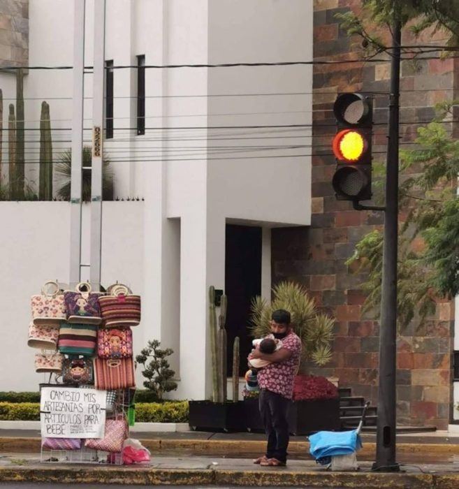 Artesano sosteniendo a su bebé en brazos y pidiendo artículos para bebé a cambio de sus artesanías 