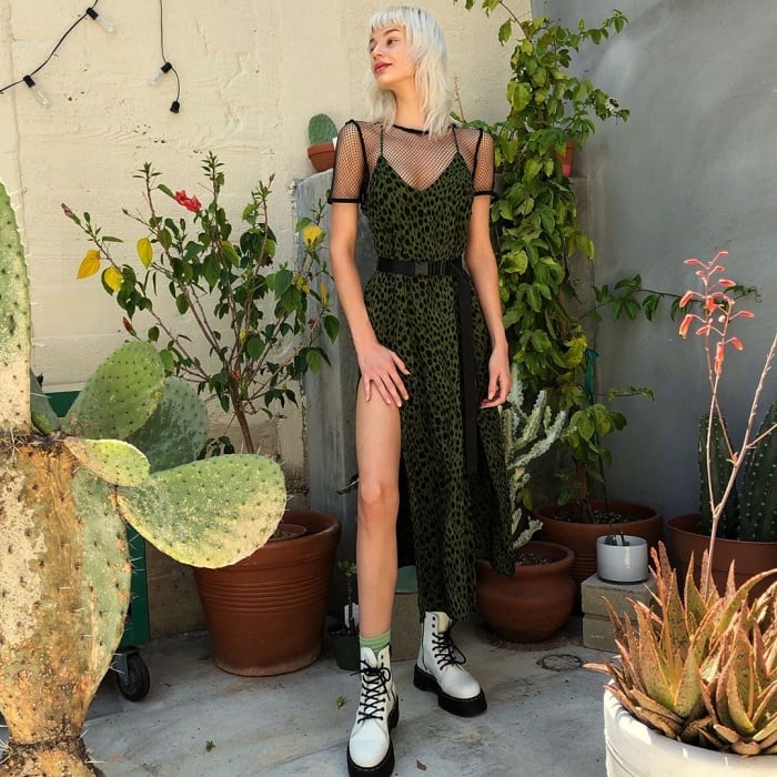 chica con cabello platinado usando un vestido verde oscuro con tul en las mangas, escote en V y abertura en la pierna, con botas de piel blancas con negro