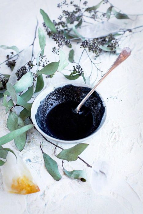 Tazón blanco con plantas verdes alrededor lleno de mascarilla de carbón activado