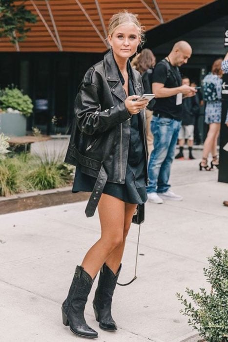 Chica posando en la calle para una fotografía mientras usa un outfit de color negro con botas vaqueras 