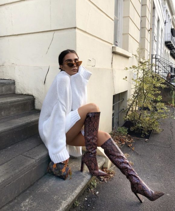 Chica sentada en unas escaleras posando para una foto mientras usa un outfit de color blanco con unas botas largas de color guinda 