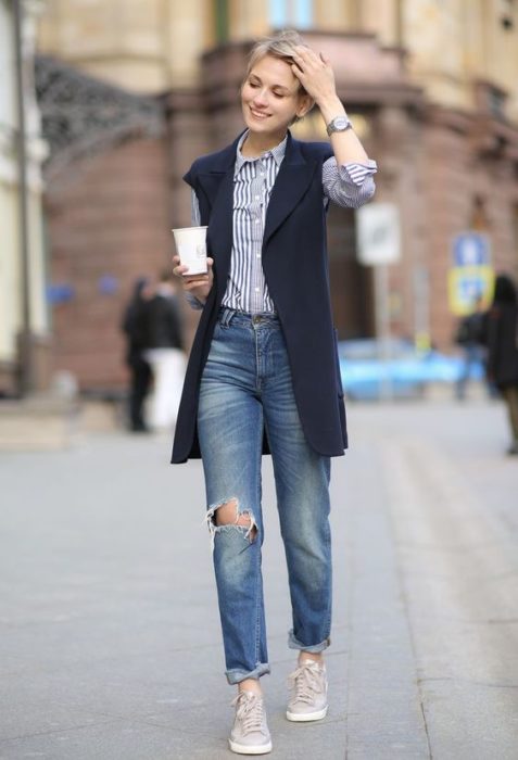 Chica usando jeans rasgados, tenis grises, camisa azul y chaleco azul marino largo