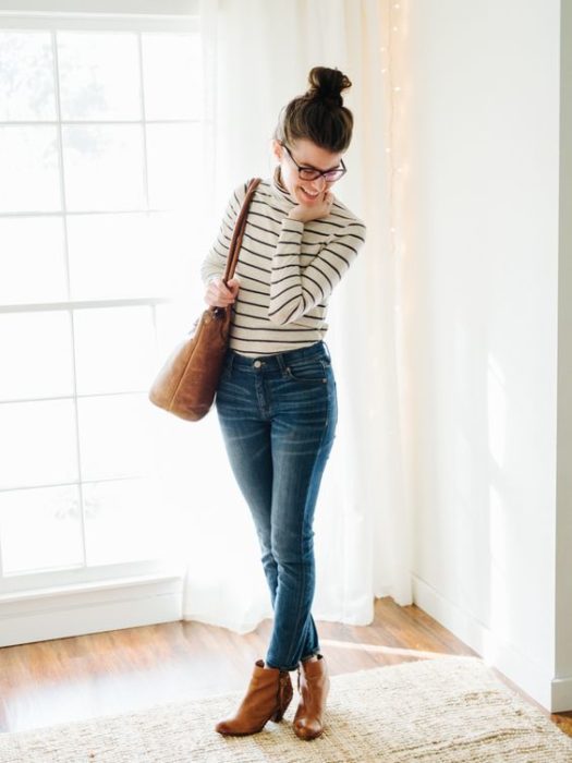 Chica con chongo y gafas con blusa blanca con rayas negras, jeans y botines cafés