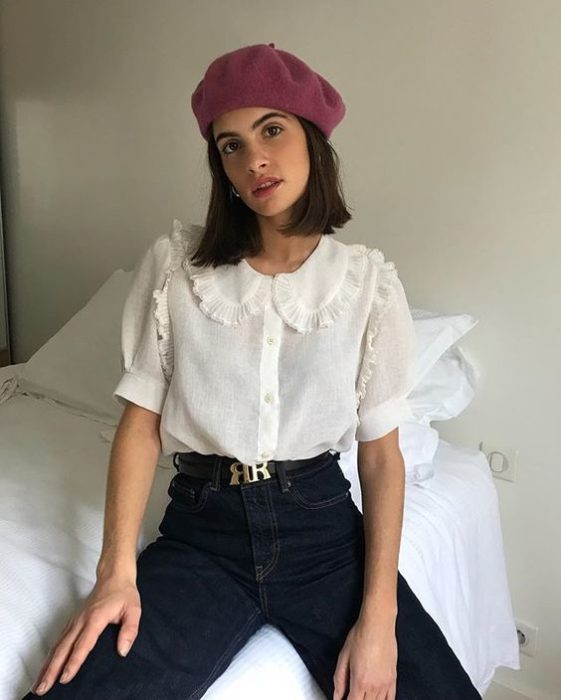 Chica delgada morena de cabello corto con gorrito rosa, pantalón negro y blusa blanca con cuello bobo