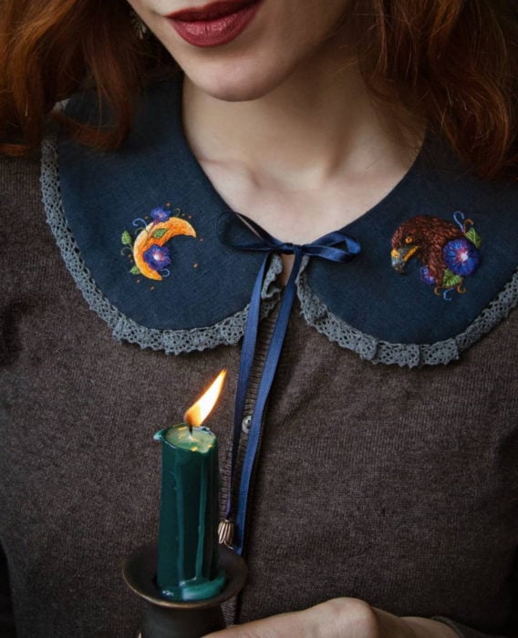 Little Pin Studio kreiert hübsche gestickte Halloween-Kragen für Kleider und Blusen.  Harry Potter, Ravenclaw, Adler, Mond und lila Blumenmuster auf marineblauem Stoff;  rothaarige Frau lächelnd hält eine grüne Kerze