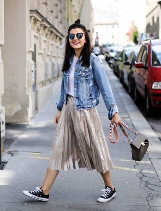 Chica caminando en la calle con falsa metálica gris, converse negros, blusa blanca y chamarra de mezclilla