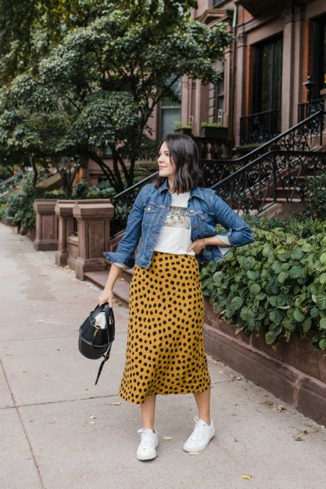 Chica de cabello corto con blusa blanca, chamarra de mezclilla y maxi falda amarilla con puntos negros y tenis blancos