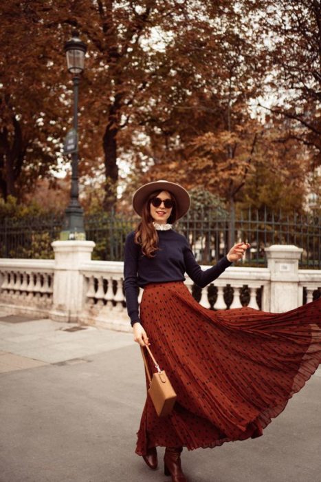 Chica de cabello largo suelto castaño con lentes de sol y sombre con suéter azul marino y maxi falda naranja 