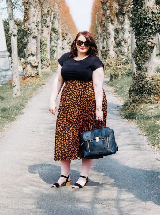 Chica plus suze pelirroja con blusa negra y maxi faldas naranja de flores y sandalias negras