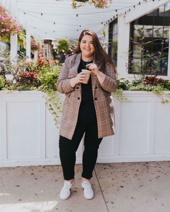 Chica curvy usando zapatos grises, con blusa y pantalón negro y blazer color arena largo