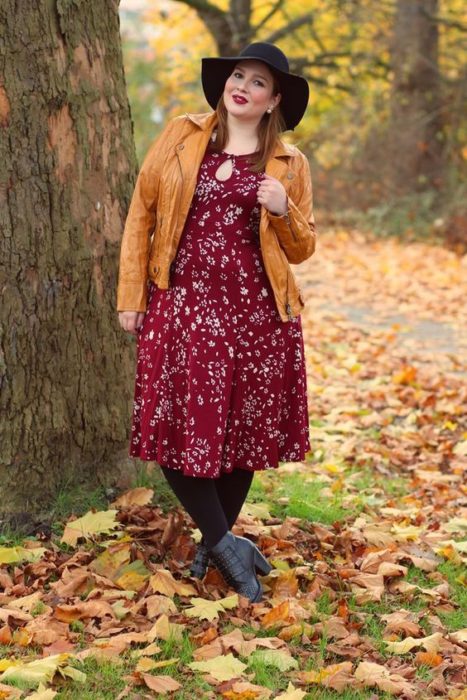 Chica curvy usando botines grises, medias y sombrero negros, vestido midi color vino y estampado de flores y chamarra color mostaza