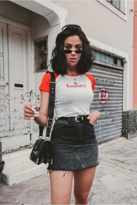 Chica morena de cabello corto con blusa gris con mangas rojas y falda negra de mezclilla 