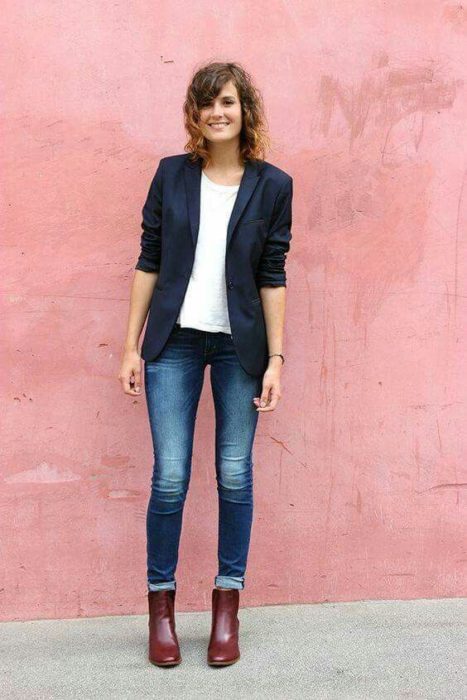 Mujer con cabello castaña corto, jeans, botines cafés, blusa blanca y saco negro